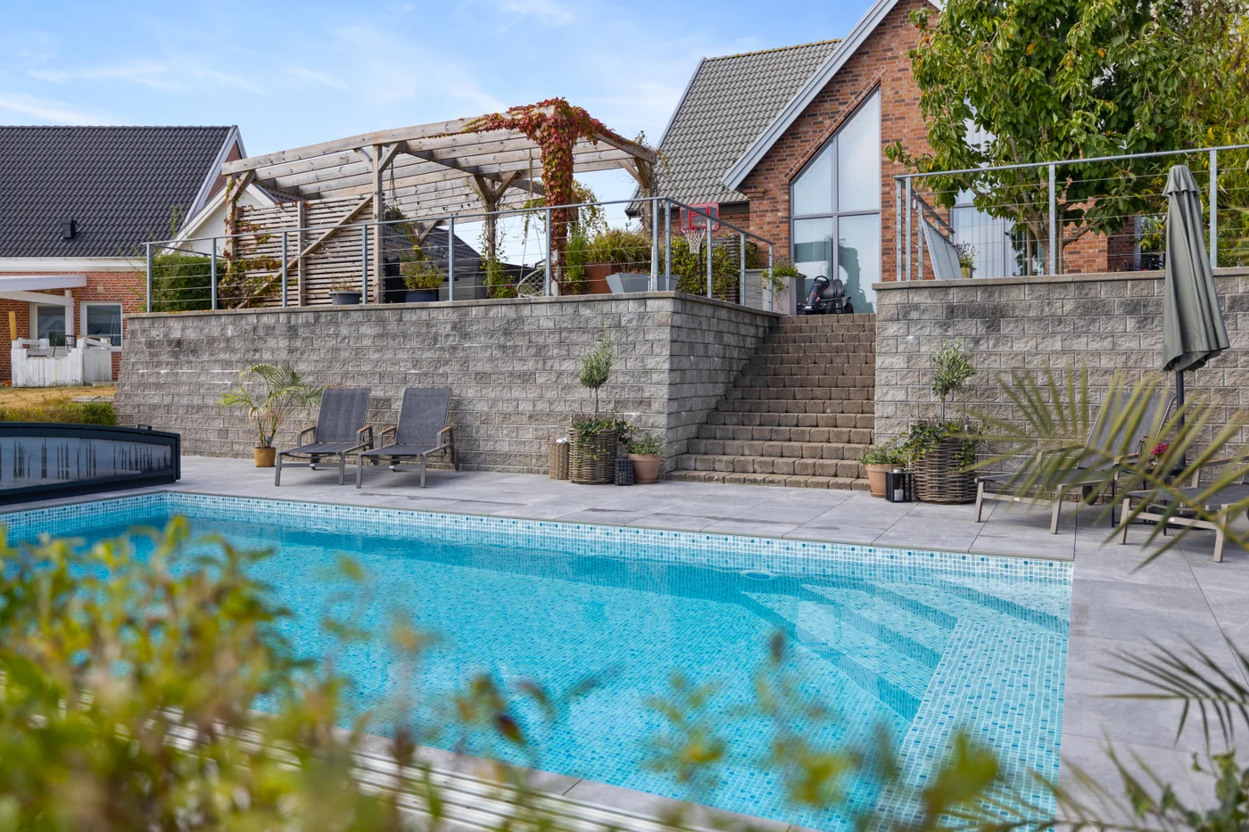 Trädgårdsutsikt och pool från kundcase Rektangulär pool med mosaikliner i trädgård med stenmur, växter och loungestolar från kundcase hos Österlens Poolcenter.